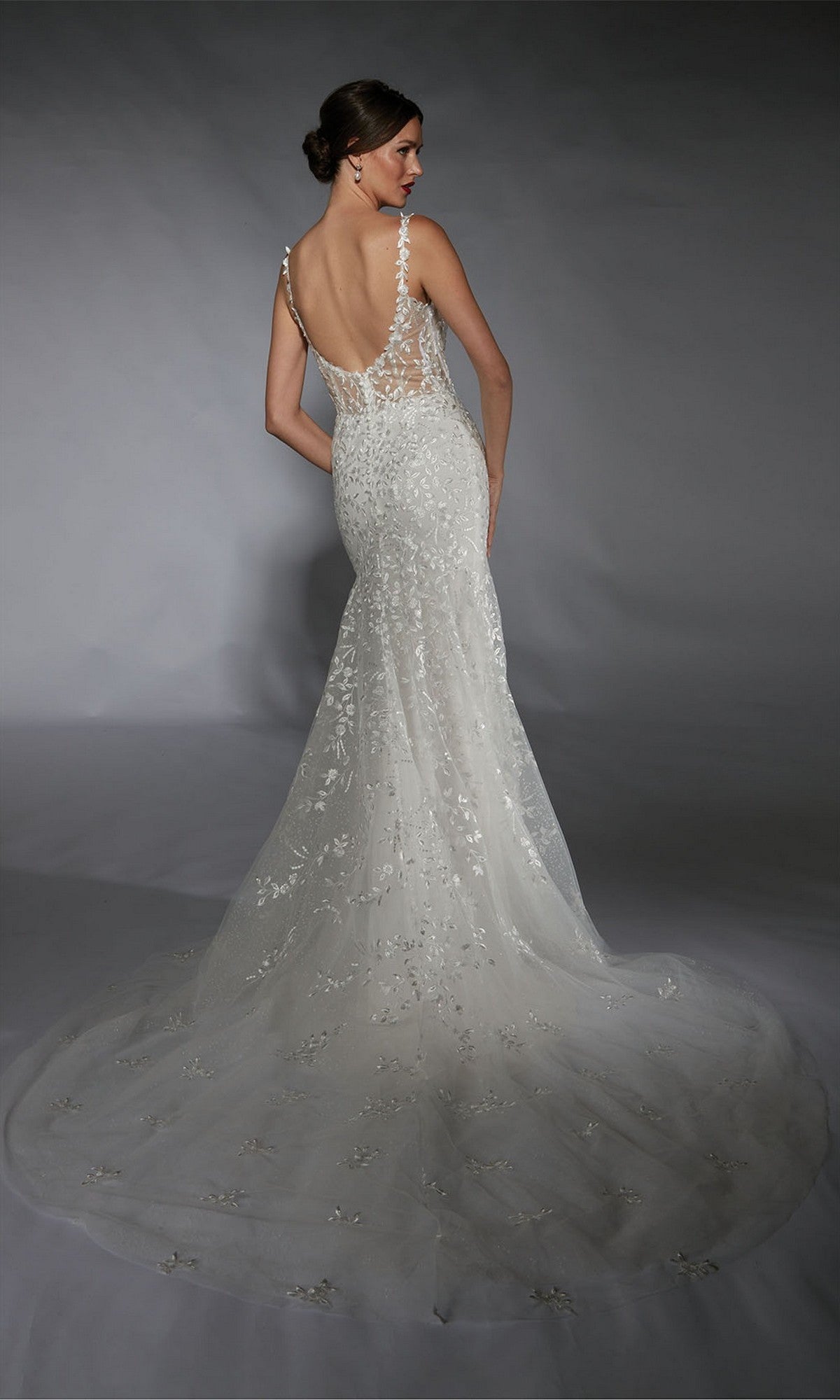 A woman wearing the Long White Dress: Alyce 7122, a backless fitted wedding gown with floral lace appliqués and a long train, stands against a neutral background facing away from the camera.